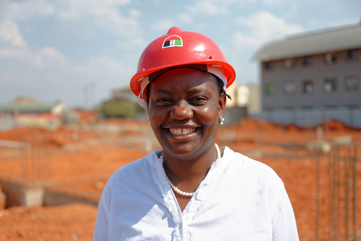 African lady with red hard hat smiling with building site behind