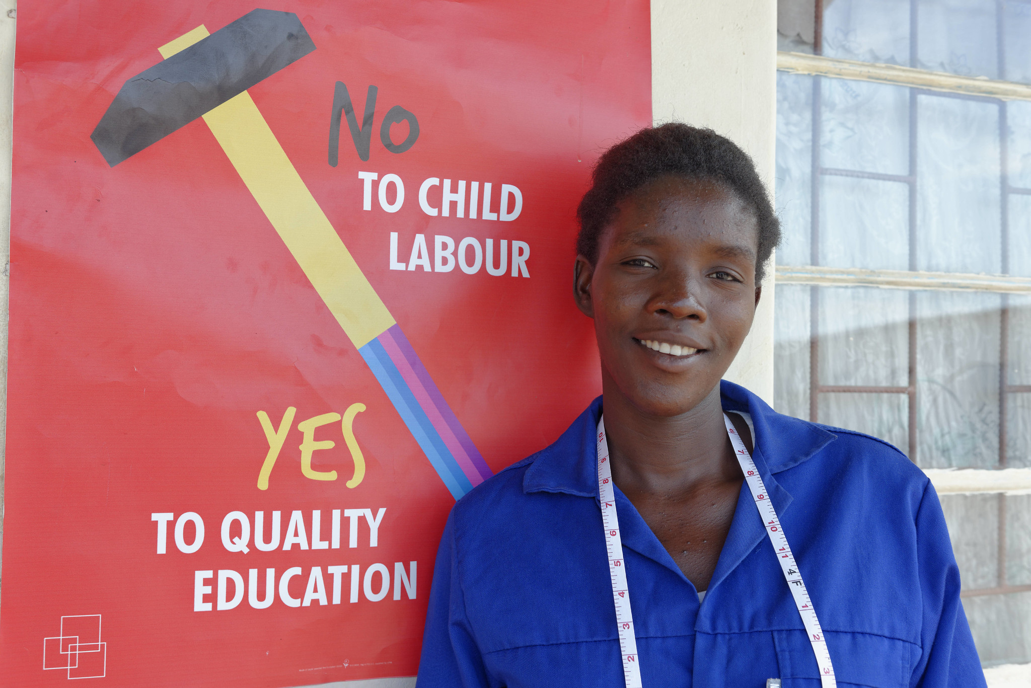 Young black lady with blue overalls and tape measure around her neck in front of an ILO Child Labour poster