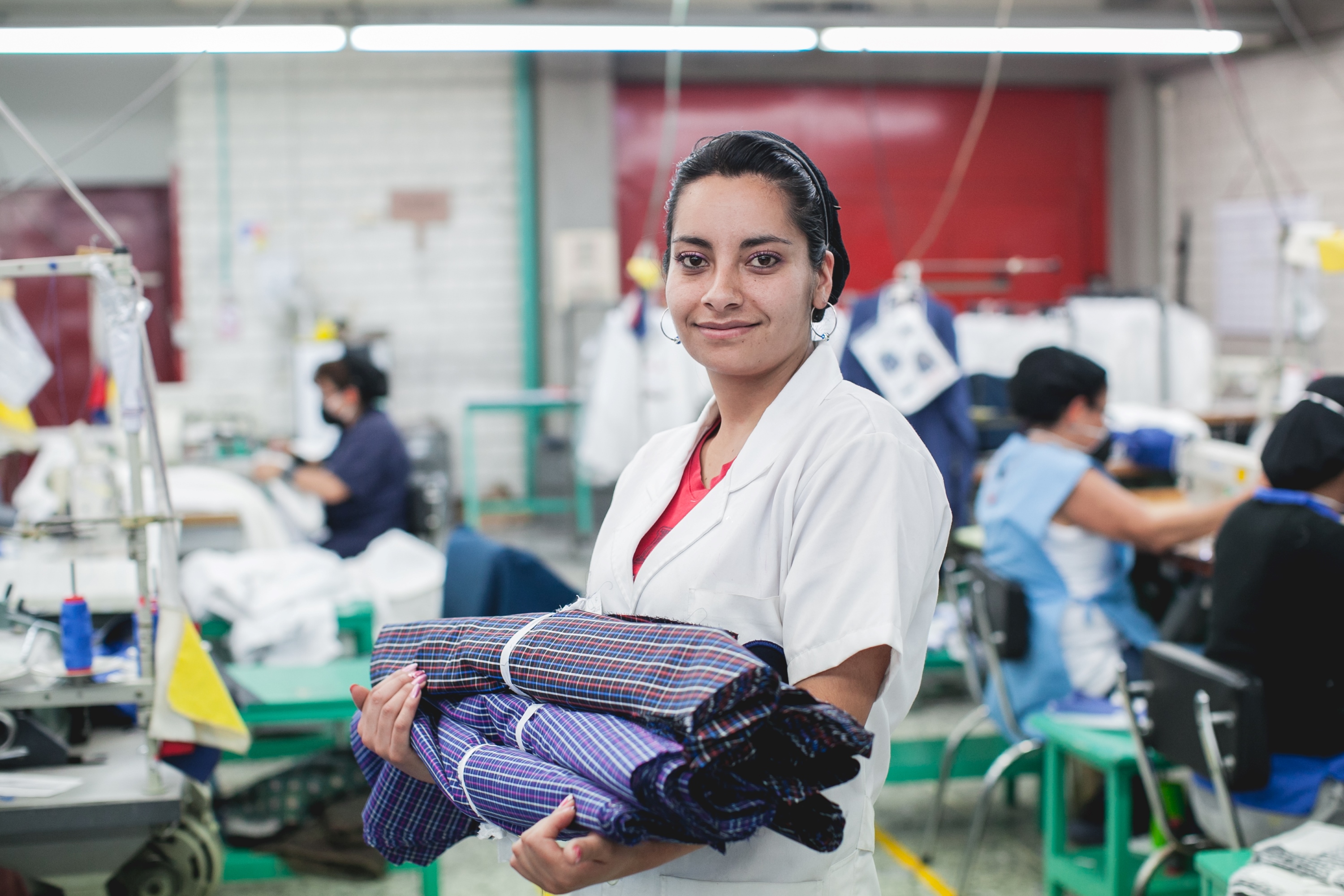 Image of young woman in fore ground holding 3 piles of material in sewing factory with other workers sewing blured behind