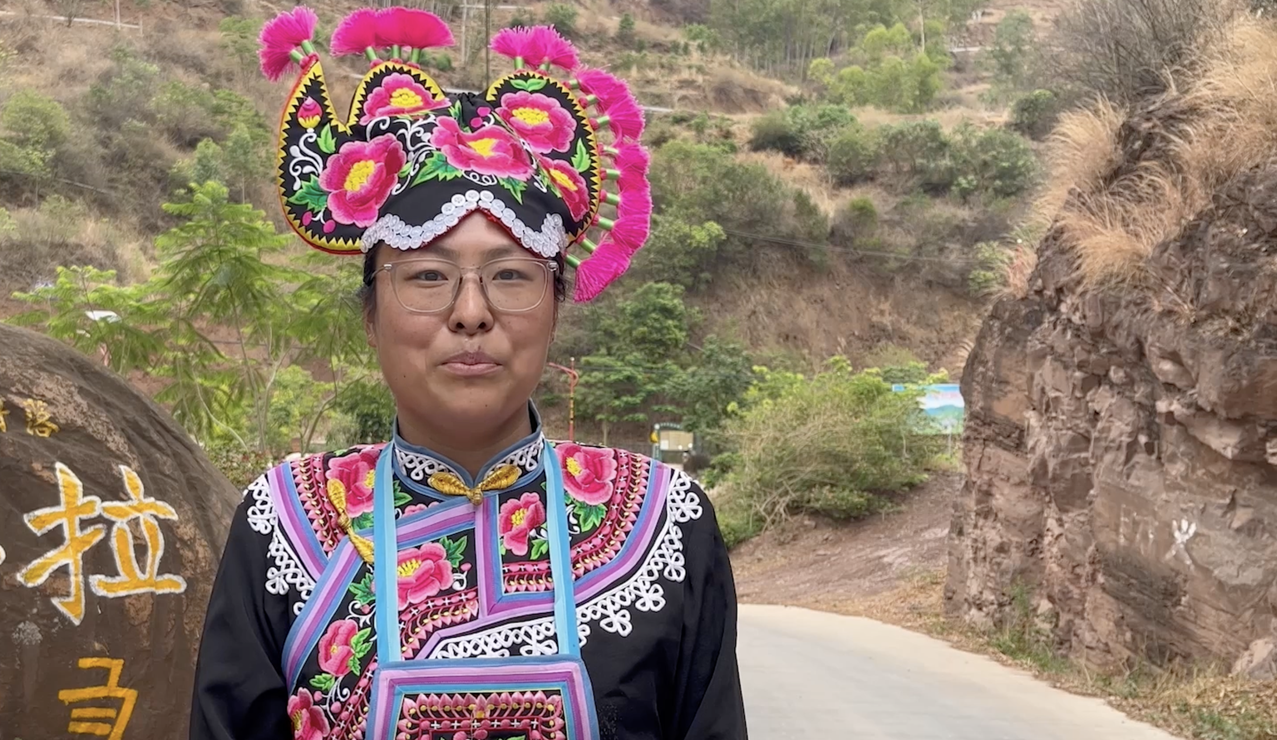 A chinese woman in a colorful outfit stands on a road by a boulder with Chinese writing on it.