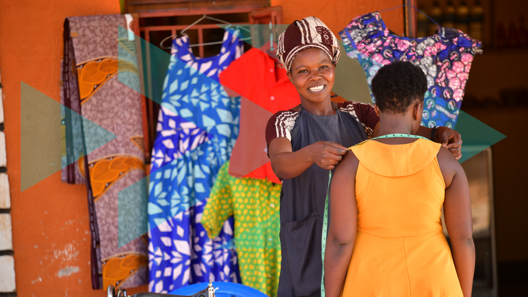 Orange brick wall with colourful dresses hanging and young black female tailor smiling and measuring another lady for a fitting