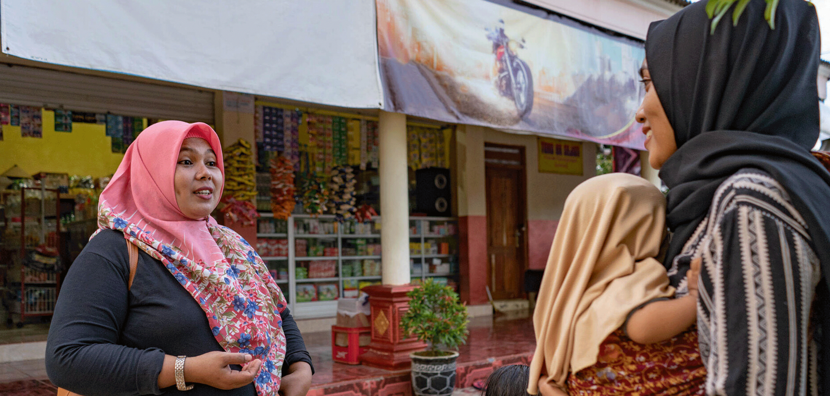 Side view of two women with headscarf, in fromt of a shop in indonedia. one woman is one carrying a baby