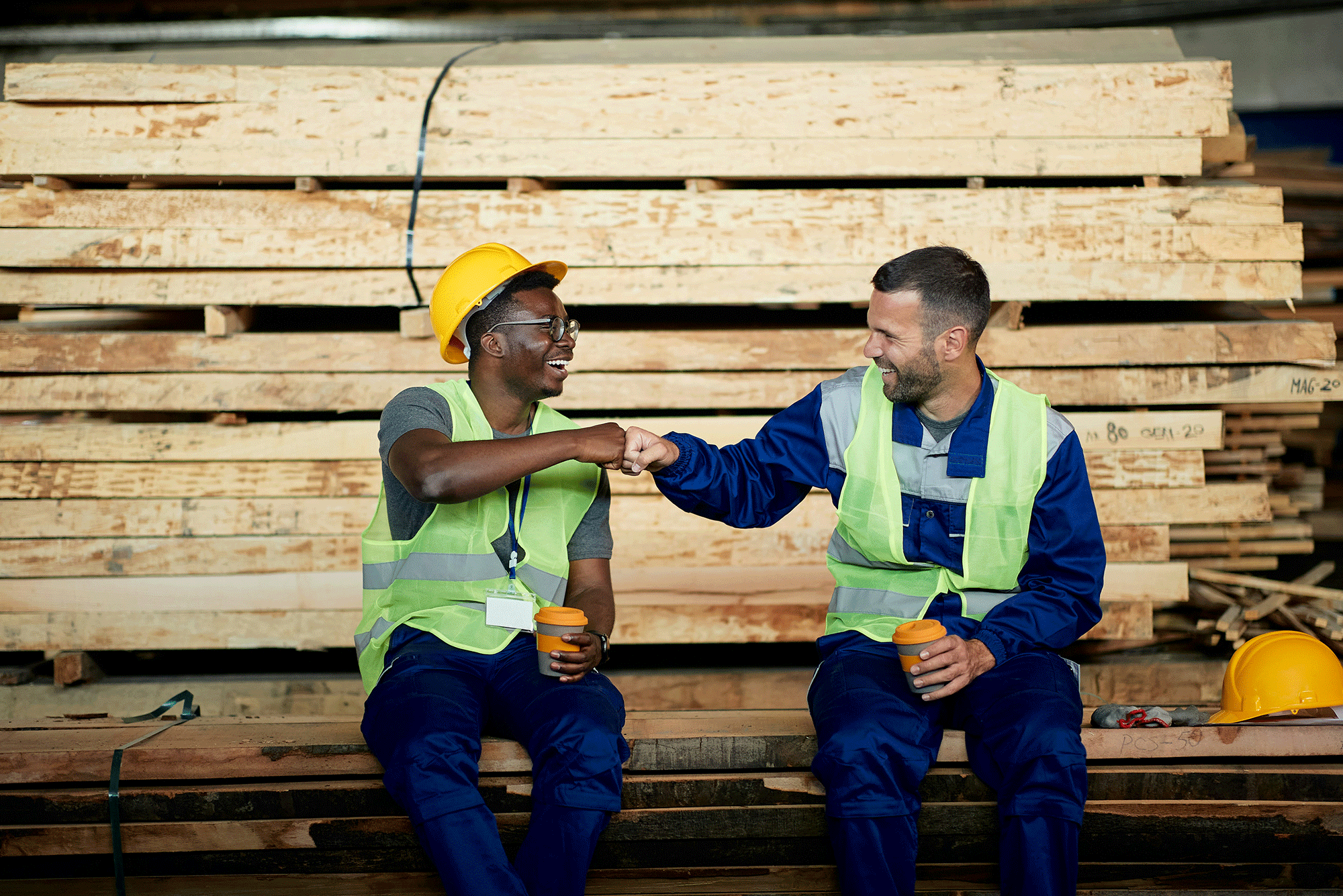 Two male workers having fun and fist bumping during their coffee break at lumber warehouse.