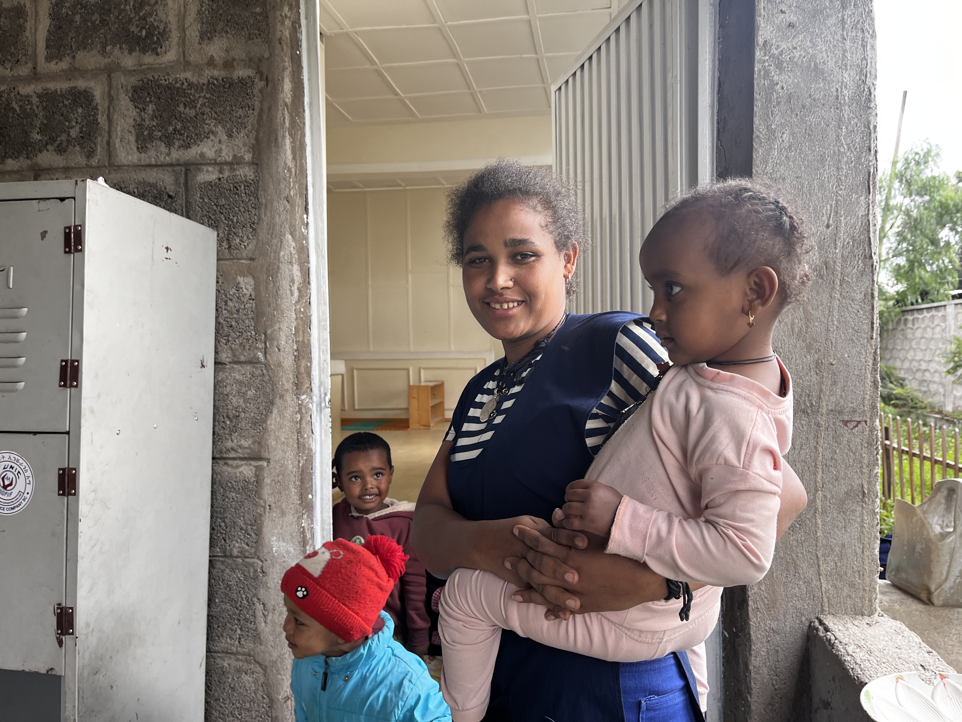 	A portrait of garment factory worker, Meseret Tamru. She stands in the doorway to a daycare centre in Addis Ababa, Ethiopia. She smiles, holding her daughter. Two other children stand nearby. (Ethiopia, 2024)