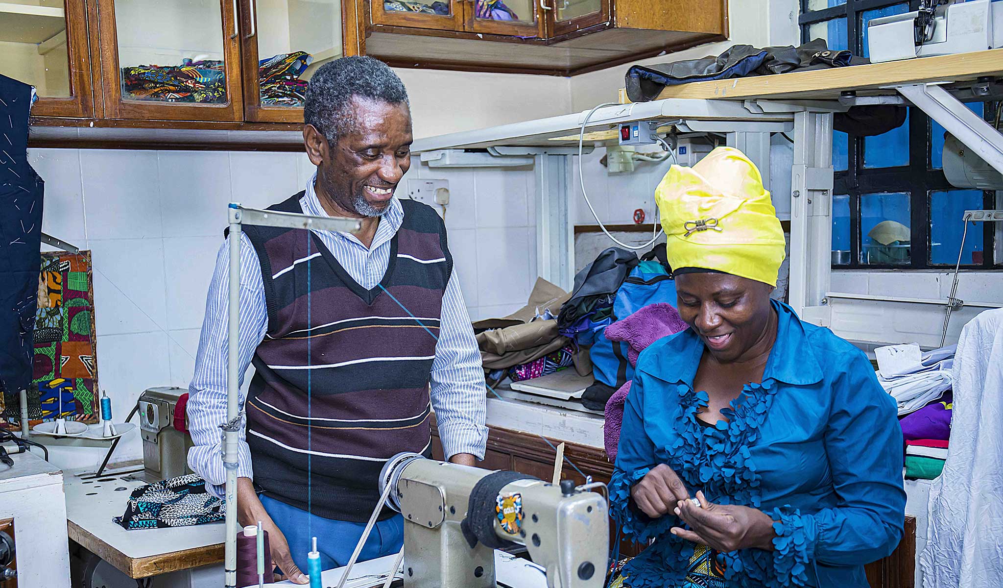 Celestine Mbaruku stands beside a trainee in a sewing workshop. The trainee and teacher both smile broadly. 