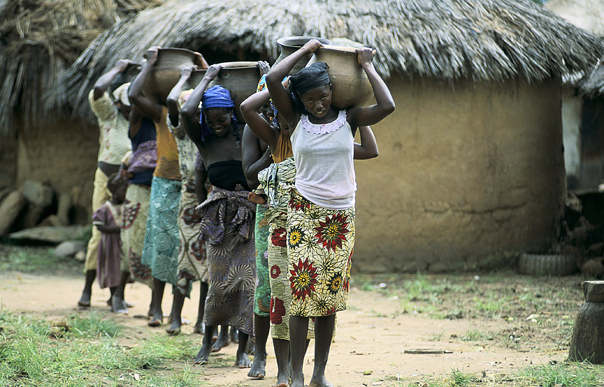 Line of black women in colourful skirts in African village carrying large round pots on their shoulders
