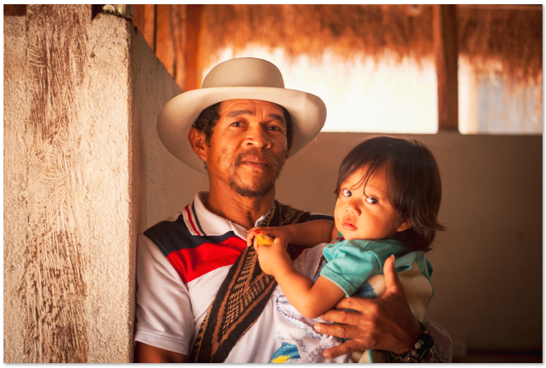 A person wearing a white wide-brimmed hat and a red, white, and black shirt stands indoors beside a textured wall, holding a young child in a turquoise shirt. Warm golden light fills the scene, with a thatched roof and softly lit background creating a cozy atmosphere. Both subjects are facing the camera.