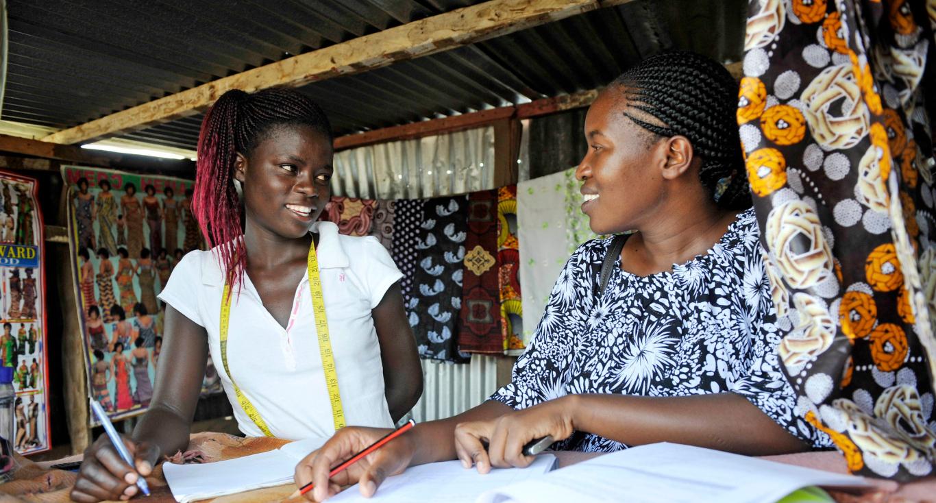 Two young black ladies talking with note pads infront of them and textile around them