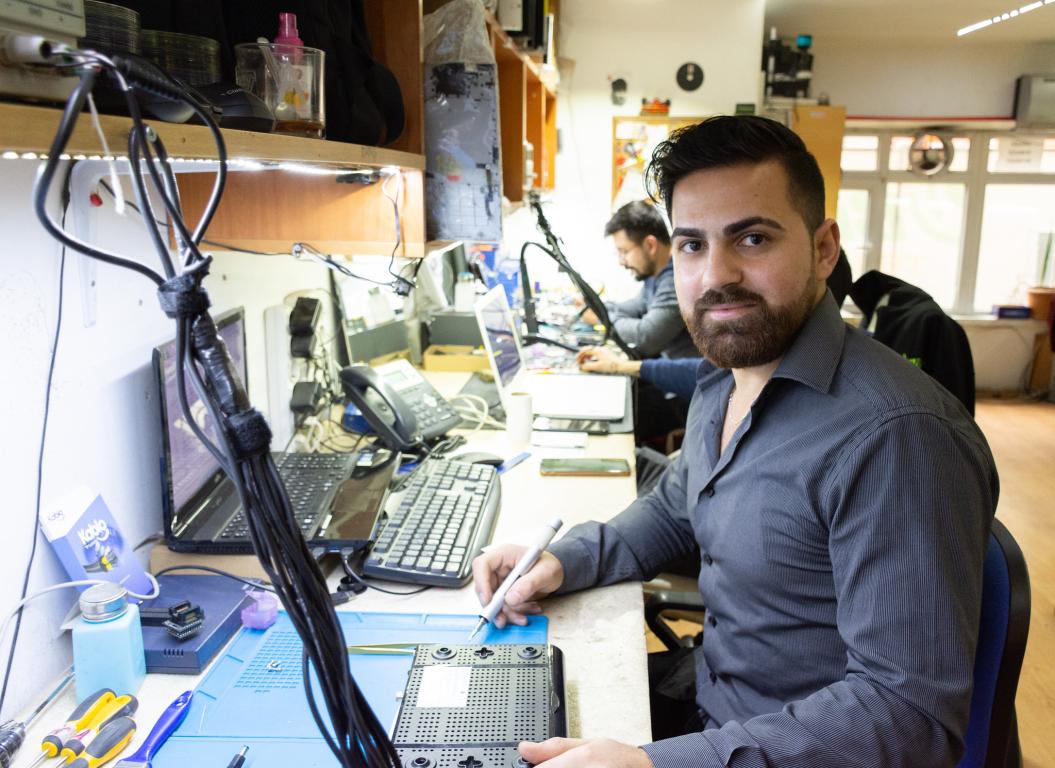 Dark haired man with beard in grey shirt sat at a desk with soldering iron