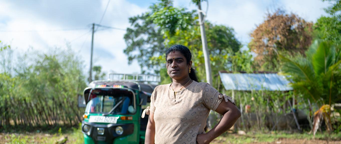 Portrait of Leena Penildas Thatkuras, standing on a path between two fields, with her hand on her hip. Her three-wheeler is parked in the background.