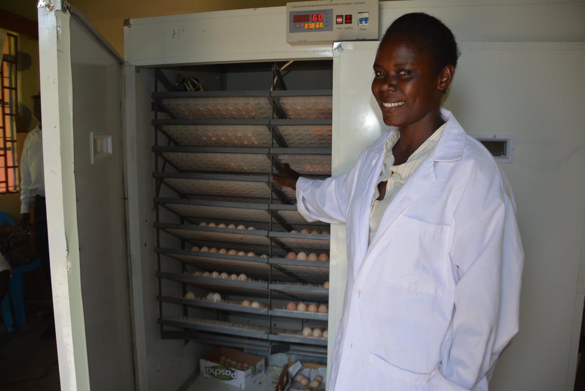 Black lady in white lab coat smiling and pointing to hatchery with eggs