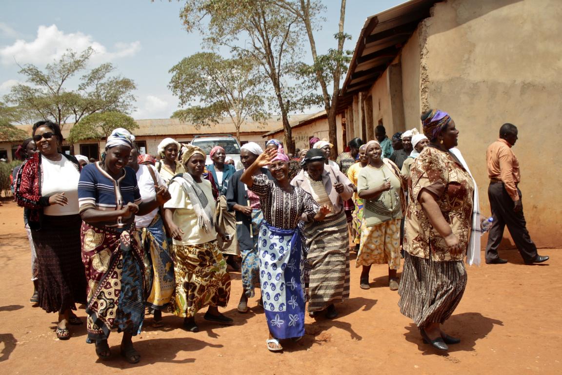 Group of black smiling women in colourful clothes in orange dirt group and low huts/buildings behind