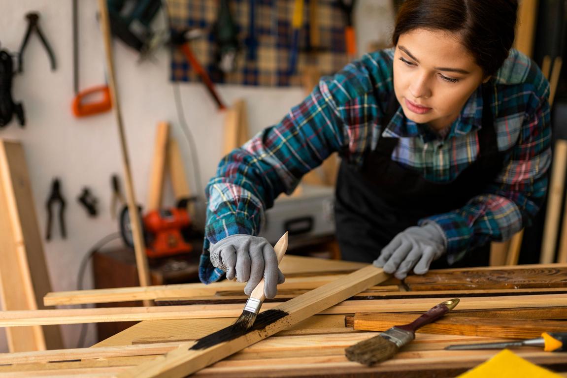 Lady in workshop with lots of wood, painting one of the wooden strips