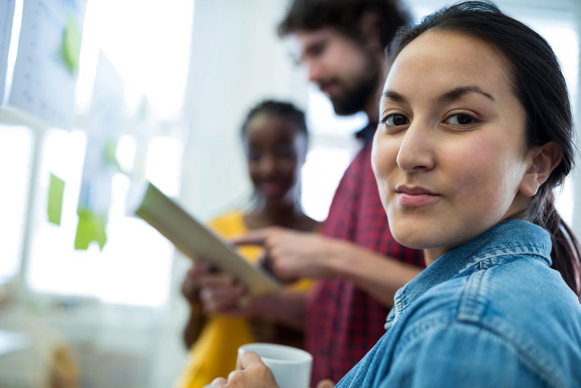young lady with blue shirt looking towards the camera with man and women out of focus behind her looking at a paper