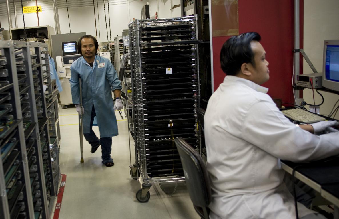 Asian man in white overalls looking at a computer with another in blue overalls walking with crutches