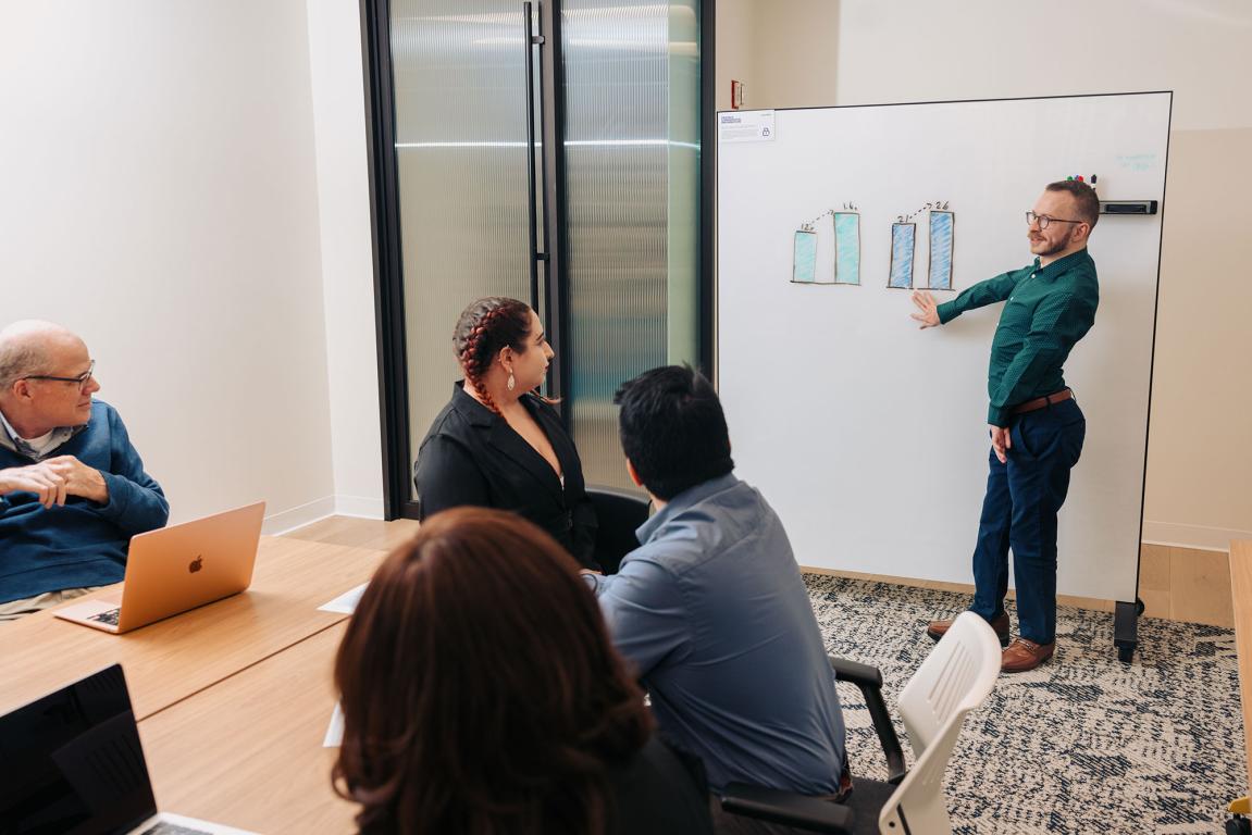 Paul presents in a conference room pointing to a white board 