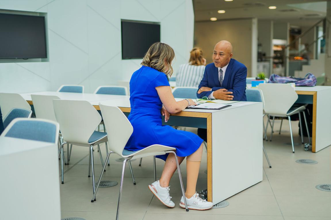 A lady in blue dress talking to a work colleague opposite each other over white table