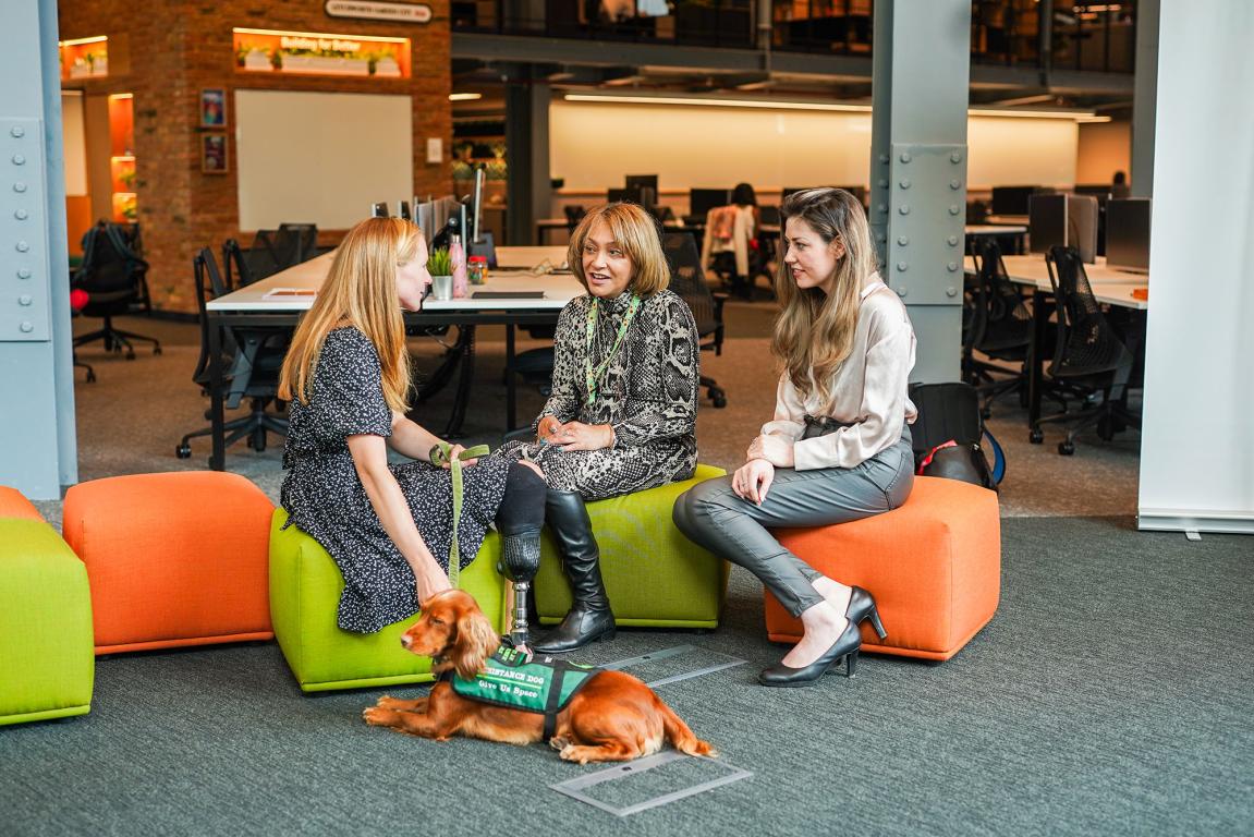 Three ladies sat in a break out area on the side of an open plan office area, one lady is stroking her guide dog