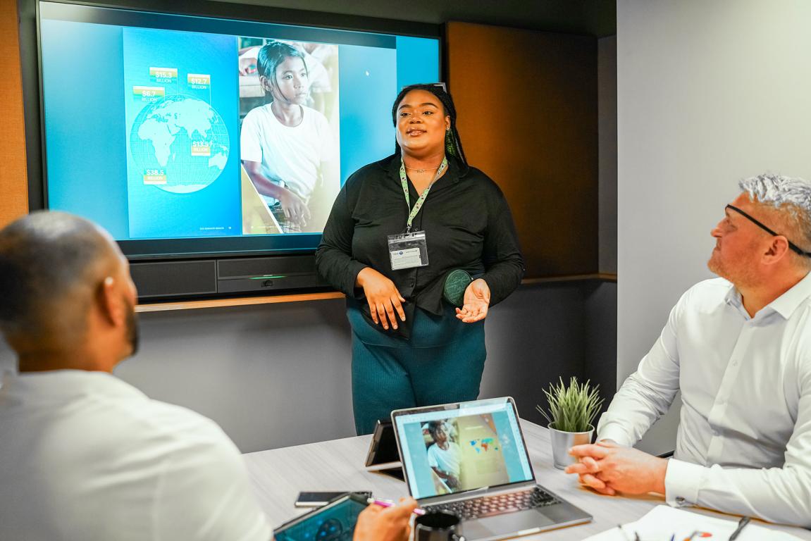Lady in front of large screen at work giving presentation to 2 male colleagues