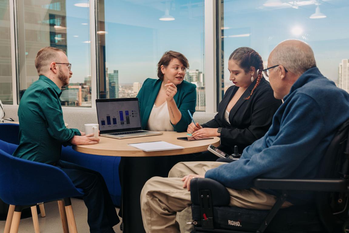 Group of four adults of various ages sat and discussing presentation on laptop around round table from office