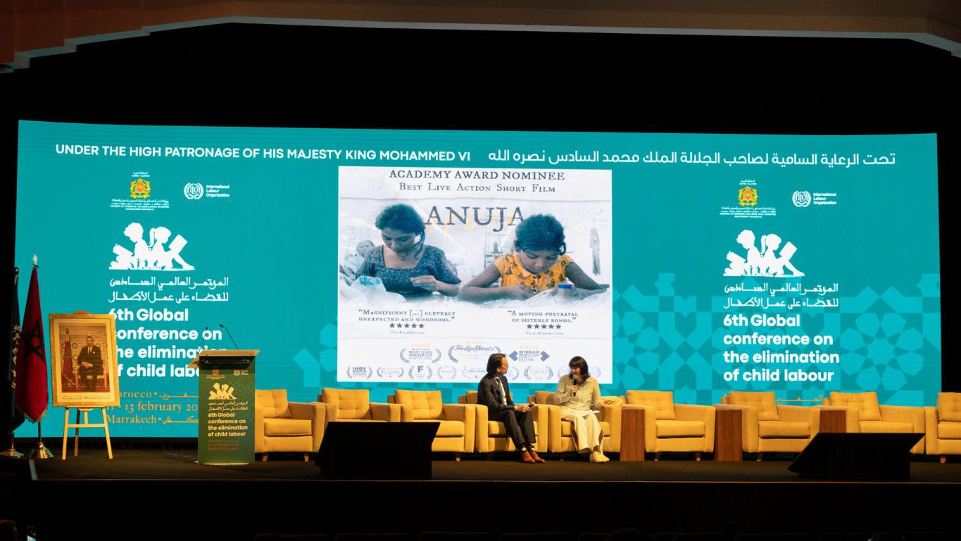 Shot of a conference room with one man and one woman sat talking infront of a promotional banner for child labour video