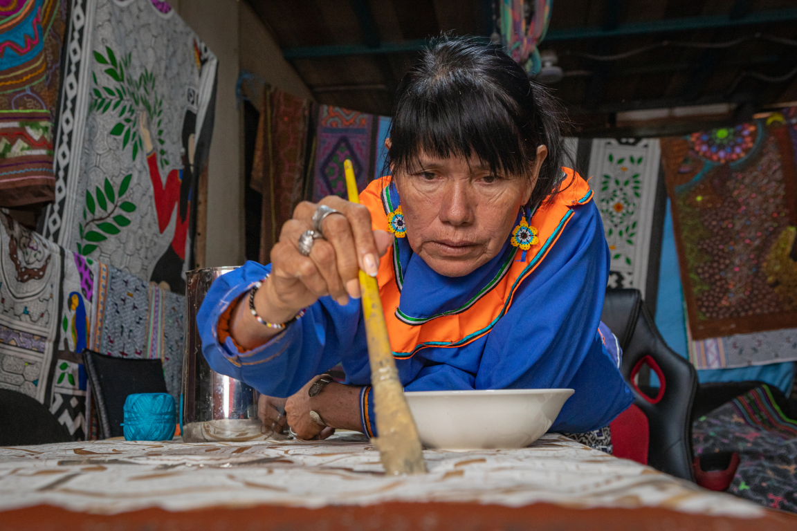 Peruvian artist with blue and orange tunic painting on cloth with many other painted pieces of material drying behind her