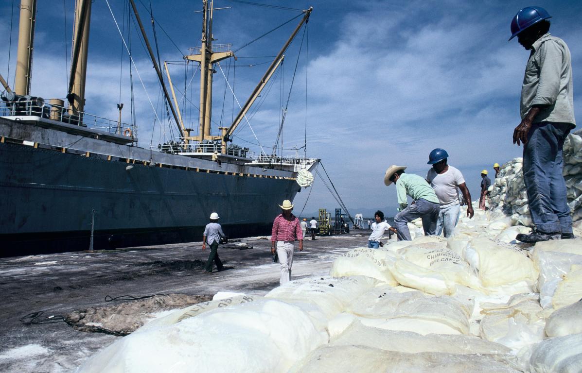 Workers standing on bags of flour in front of a small cargo ship at a dock