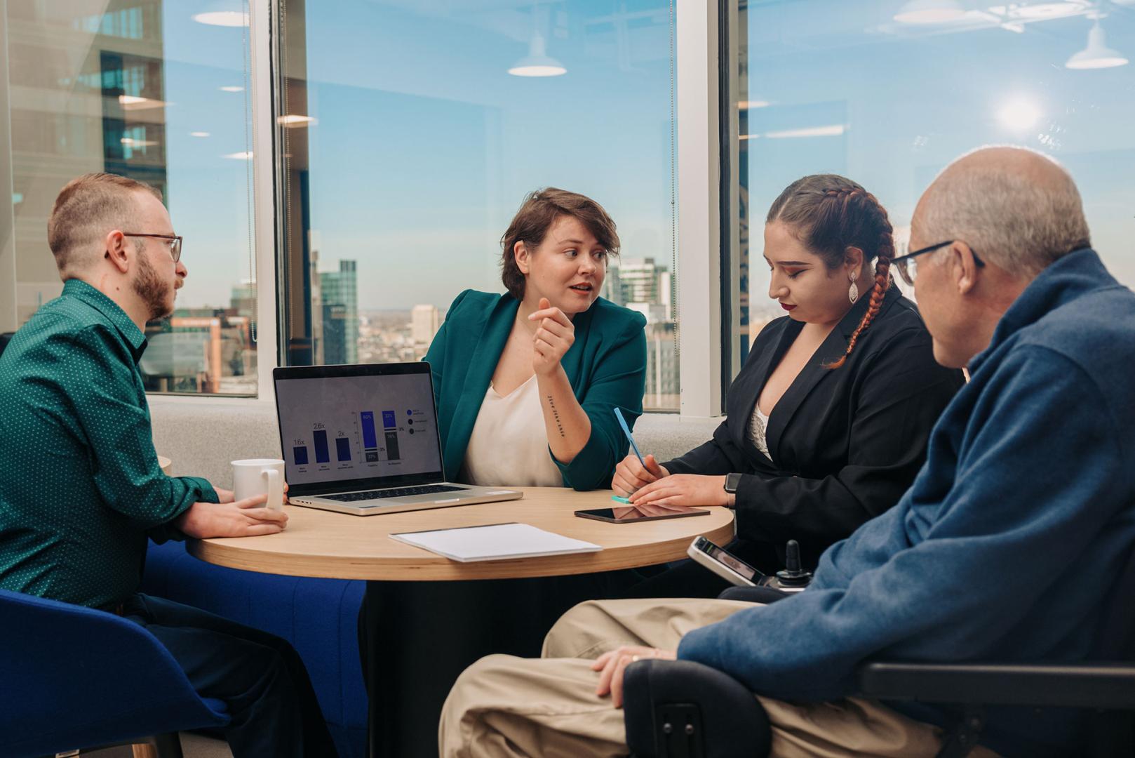 Group of four adults of various ages sat and discussing presentation on laptop around round table from office